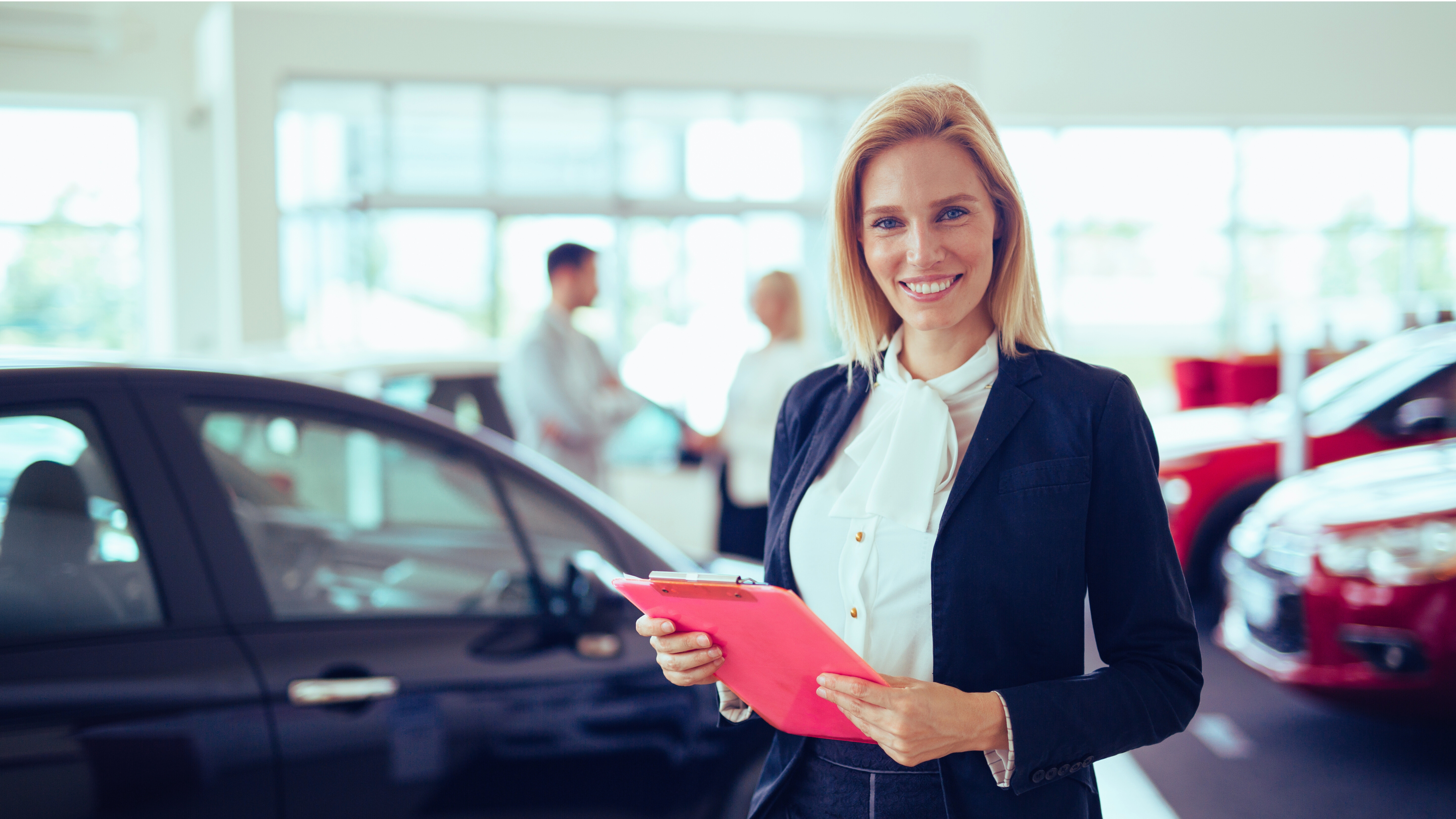 Blonde Verkäuferin steht lächeln vor einem Fahrzeug im Autohaus